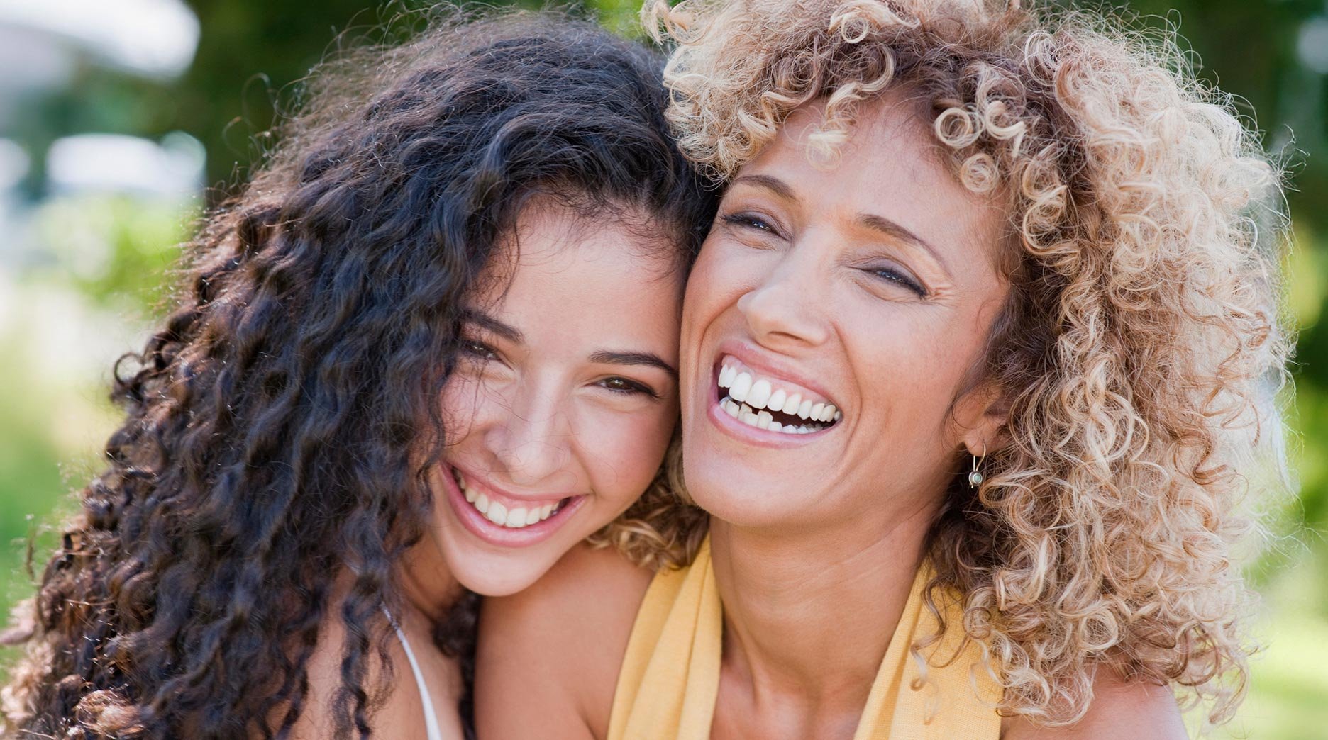 Close up of two female best friends laughing and embracing each other. They each have beautiful curly hair, one has long brunette 2C curls, while the other fashions shoulder length, blonde, 3B curls.